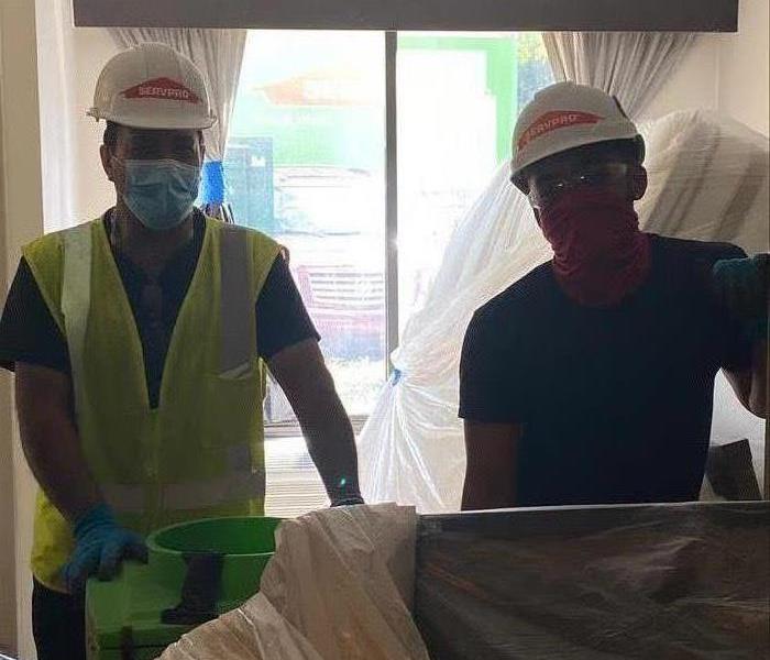 Two SERVPRO employees standing in a room next to furniture covered in plastic.